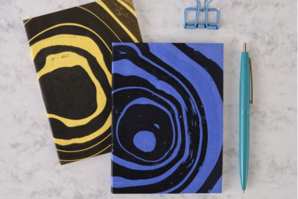 Two notebooks, one blue and one yellow with a circular black design are on a marble countertop with a blue bulldog clip above and a blue pen to the right