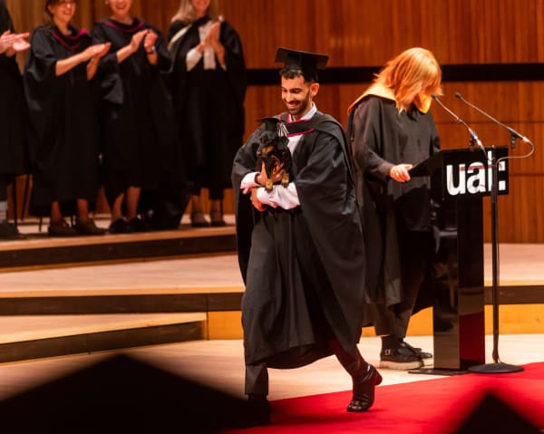 Graduate walks across stage holding a dachshund wearing a small graduation hat