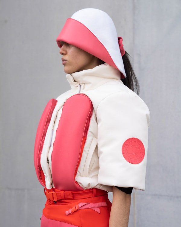 A side shot of a model in functional outdoorwear in pink and white including a sculptural hat
