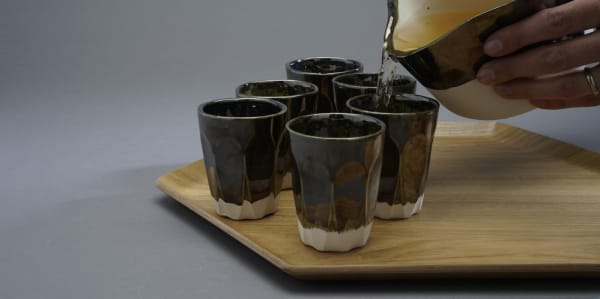 set of six brown ceramics cups on an hexagonal wooden tray. A hand is pouring tea from a ceramic jug into one of the cups