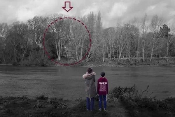 A photograph of two people standing on the bank of a pond looking across the water to some trees. A red dotted line circle and red arrow point towards one area of the scene. 