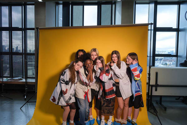 A group of students stand for a photoshoot with a bright yellow background they are giggling and smiling 