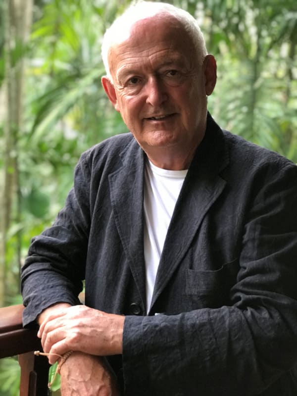 James Acheson leans against a railing in front of some trees. He has short, white hair and wears a white t-shirt underneath a dar jacket. 