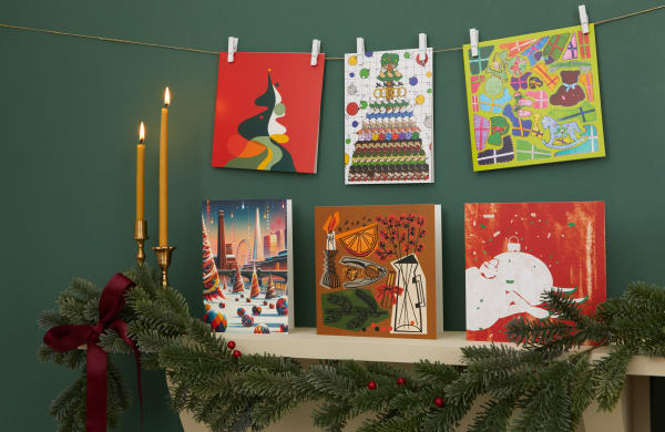 A festive holiday scene featuring a collection of colourful Christmas cards displayed against a dark green wall. Three cards hang from a string with clothespins above: an abstract Christmas tree design on red, a grid of toy soldiers and baubles, and a bright patchwork illustration with gifts and rocking horses. Below, more cards are propped on a mantel: a vibrant cityscape with Christmas trees and the London skyline, a warm-toned still life with a candle, orange slices, and a vase, and a red-and-white card with an abstract figure. The mantel is decorated with evergreen garlands, red berries, a dark red ribbon, and two lit gold candlesticks.