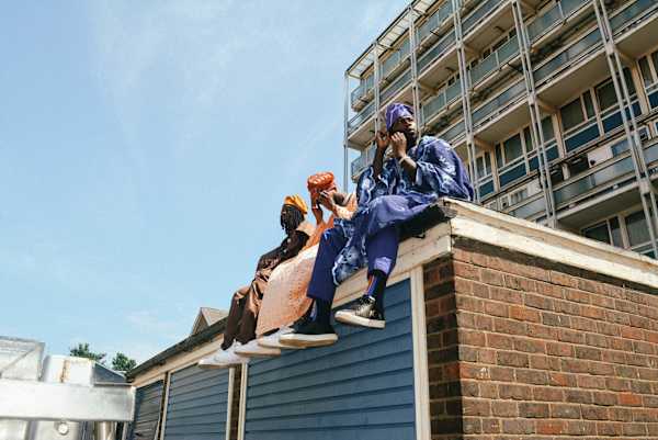 Three people wearing brightly patterned traditional clothing sit on the edge of a rooftop against a backdrop of urban buildings and a clear blue sky.