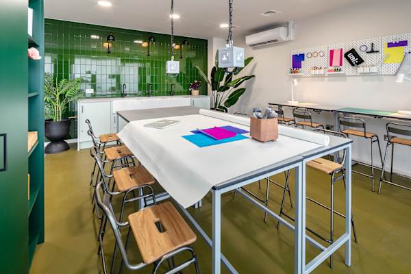 Creative space with a large white-topped work table covered in paper and bright coloured card, surrounded by wooden stools. Hanging plug sockets sit above the table, with a green tiled wall and Belfast-style sinks behind, and pegboards of art supplies on the right.
