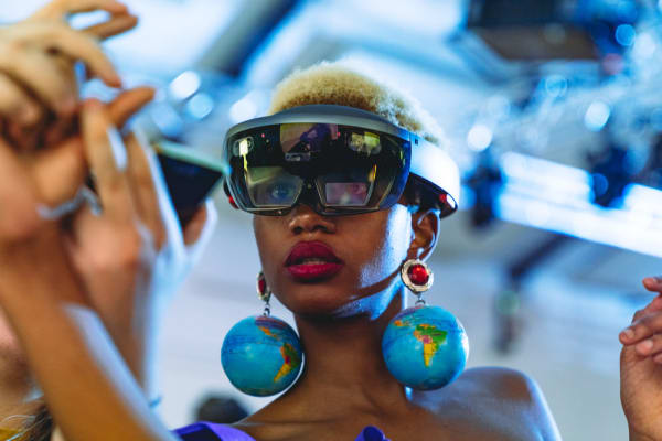 Woman wearing VR glasses and large, dangling globe earrings. We are looking upwards at her head and shoulders 