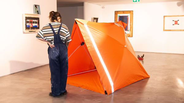 nt like structure in the centre of the gallery surrounded by various different size abstract white, blue and red and orange paintings on the walls. There is a girl looking at the tent dressed in blue dungarees.
