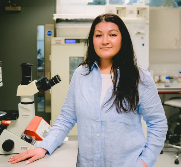 A woman in a blue shirt faces the camera and leans against a desk next to a microscope.
