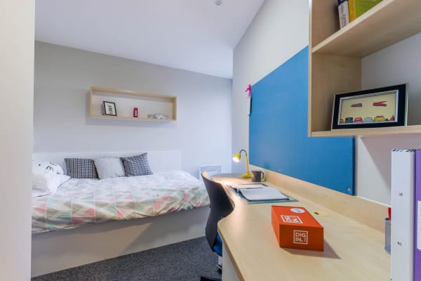 Standard room view of 3/4 bed, long desk with overhead shelves and pinboard.