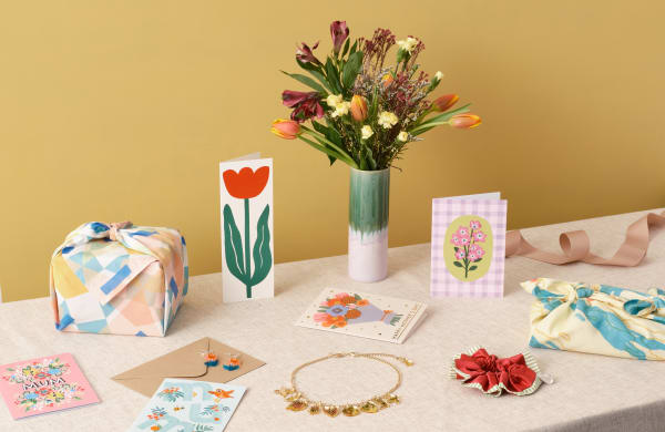 A display of colourful gifts and cards arranged on a beige table against a mustard-yellow background. The items include a wrapped gift in pastel geometric fabric, a tall green-and-pink vase with a bouquet of fresh flowers, handmade greeting cards featuring floral designs, a pair of orange and blue flower earrings, a gold charm bracelet, a red and green ruffled scrunchie, and another wrapped package in marbled yellow and blue fabric. A peach satin ribbon is loosely coiled on the right side of the table.