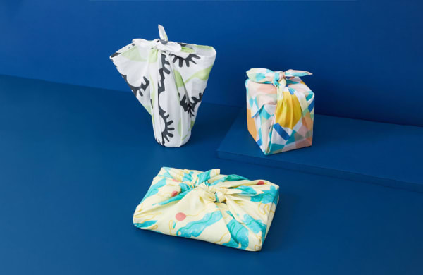 Three gifts wrapped using the Japanese furoshiki technique are displayed against a vibrant blue background. Each gift is wrapped in a different colourful patterned fabric: one with a bold black and green abstract design, another with pastel geometric shapes in peach, blue, and yellow, and the third with a flowing aqua and yellow marbled pattern. The fabric is knotted neatly on top of each package, showcasing an eco-friendly and stylish approach to gift wrapping.