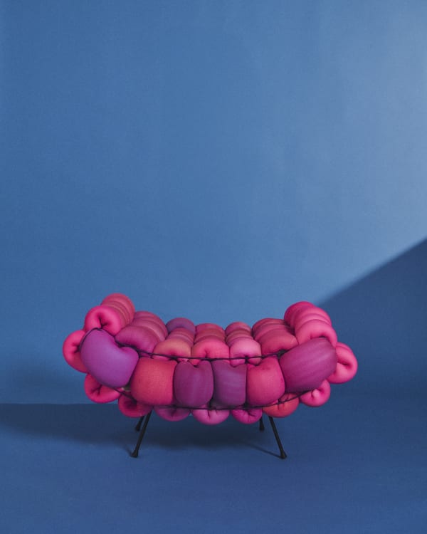 A padded, bobble design pink and red backless seat with 2 legs and an upward curve, against a blue blackground.