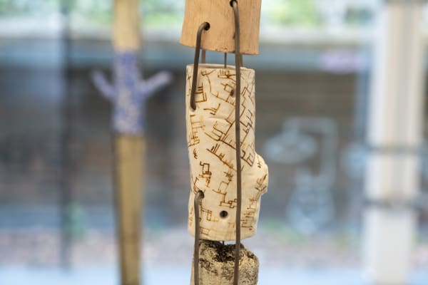 Close-up of an abstract sculpture with intricate, maze-like patterns on a white, cylindrical piece threaded through metal rods, set against a blurred background.