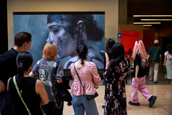 Students walking past and watching a big screen.