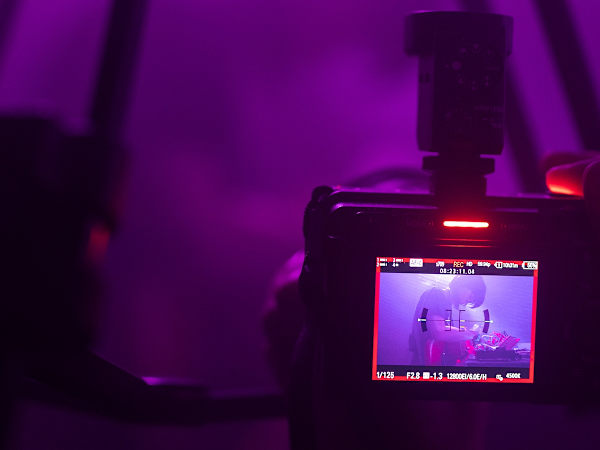 Camera filming a performance in a studio lit with purple light.