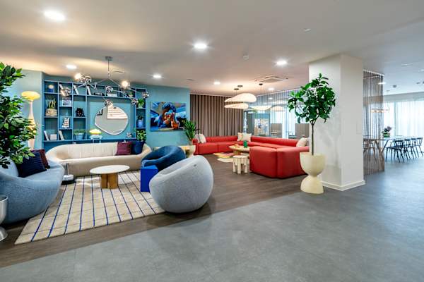 Open-plan lounge with pale blue and cream sofas, round grey and navy armchairs and a blue-lined rug, in front of a blue shelving unit with a mirror and artwork. A large coral corner sofa sits beyond, with a dining area, plants and pendant lights in the background.