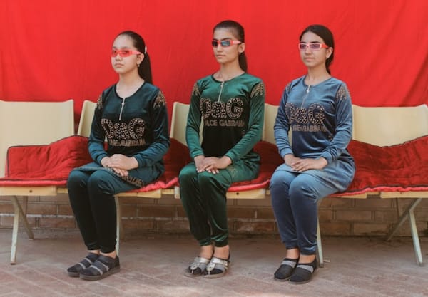 three models with hair pulled back into ponytails sit on chairs draped with red velvety material, all wearing red sunglasses and dolce & gabbana print garments.