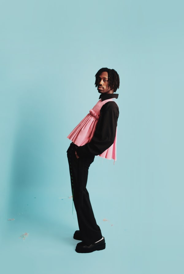 Male model posing in front of a baby blue backdrop wearing a black pinstripe shirt and trousers with a dusty pink ruffle top