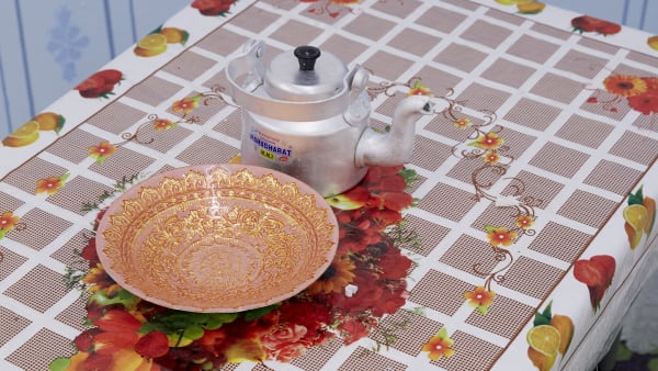 Table with decorative tablecloth with grid pattern and fruit. On the table is a metal teapot and decorative bowl. and 