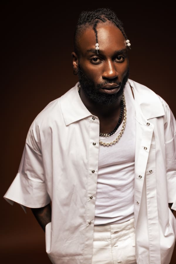 Kojey Radical stands in front of a dark background. Khojey has short, braided hair and wears a white vest, white shirt and white trousers. 