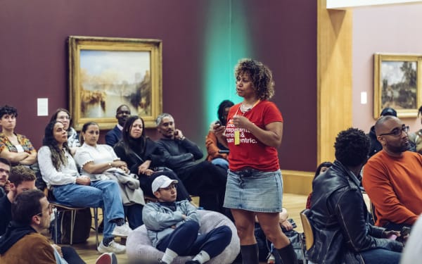 A group of people sitting on chairs and beanbags in a gallery space. One person standing in the middle speaking.