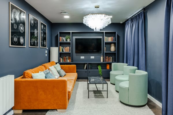 View of a common room featuring a cozy sofa area in front of a large flat-screen TV, library-inspired decorations and an statement chandelier.