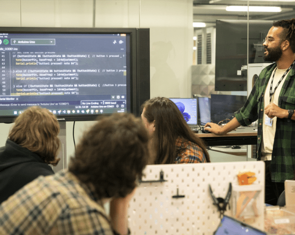A tutor is talking to a group of people about coding displayed on a screen in front of them