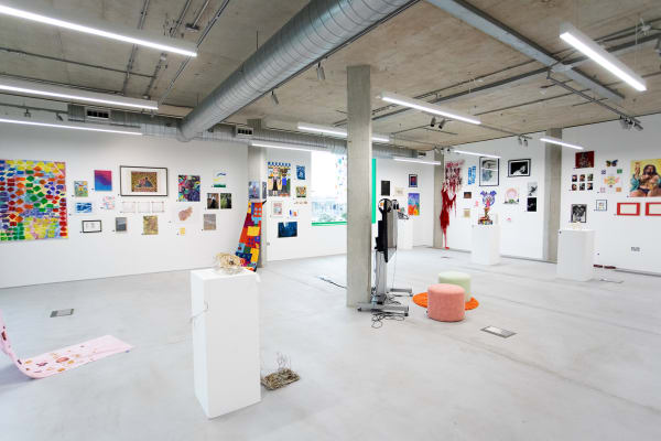 A contemporary art gallery with white walls displays various colorful artworks and sculptures; pedestals and stools are scattered around the spacious, well-lit room.