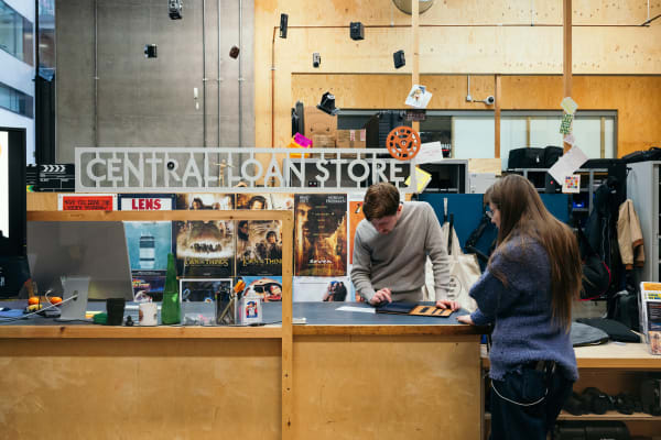 The Central Loan Store helpdesk. A sign above says Central Loan Store in bold capital lettering. There are posters hanging and there is a large Mac screen visible. A member of staff is behind the counter and another is on the other side facing them. On the countertop is a tablet and one of the staff members is swiping the screen.