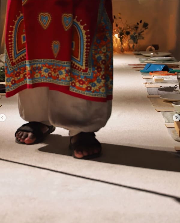 A person in sandals and a long red tunic and white undergarment is standing on a table set to the right hand side with cutlery and other materials. Flowers and soft lights are visible at the far end.