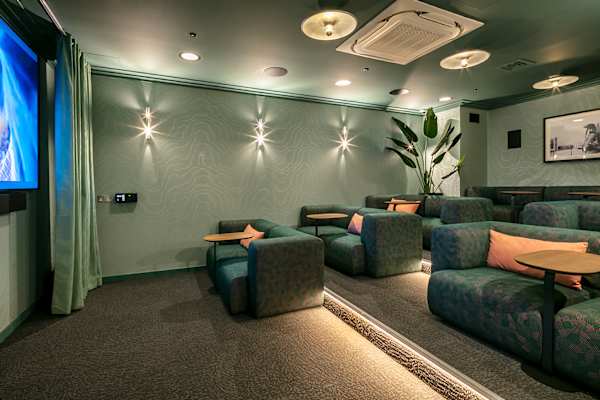 Cinema room at Archwood House with tiered rows of green patterned sofas, each with a small wooden side table and coral cushions. A large screen sits to the left beside a pale green curtain, with wall lights, a tall plant and a framed black-and-white photograph against sage green walls.