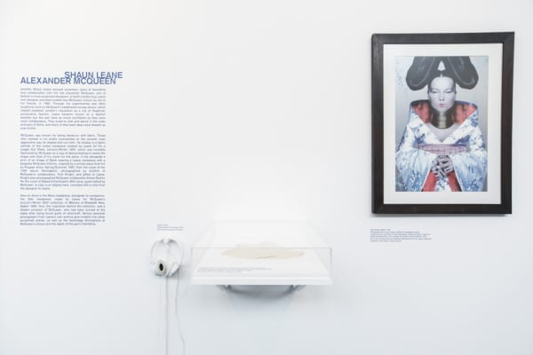 on the right side a framed portrait of a model in a silver and red kimono influence style garment, with elongated choker and hair styled in large disks. in the middle an interactive display with headphones. on the left a block of text titled 'Shaun Leane / Alexander McQueen'.
