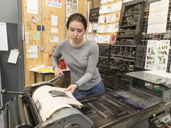 A student stands at a printing press, creating a black and white typographical print. They are surrounded with other print samples and letter press artefacts. 