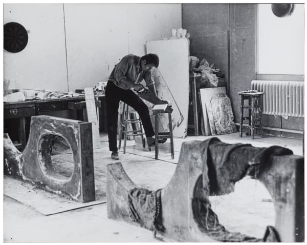 Black and white photograph of a man sawing a plank of wood in a sculpture studio, with abstract sculptures in the foreground