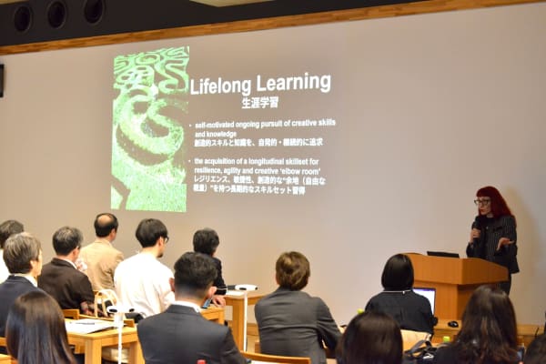 Dr Marenko presenting at a podium in front of an audience, with a slide on screen and the text 'Lifelong Learning' and description in English and Japanese. 