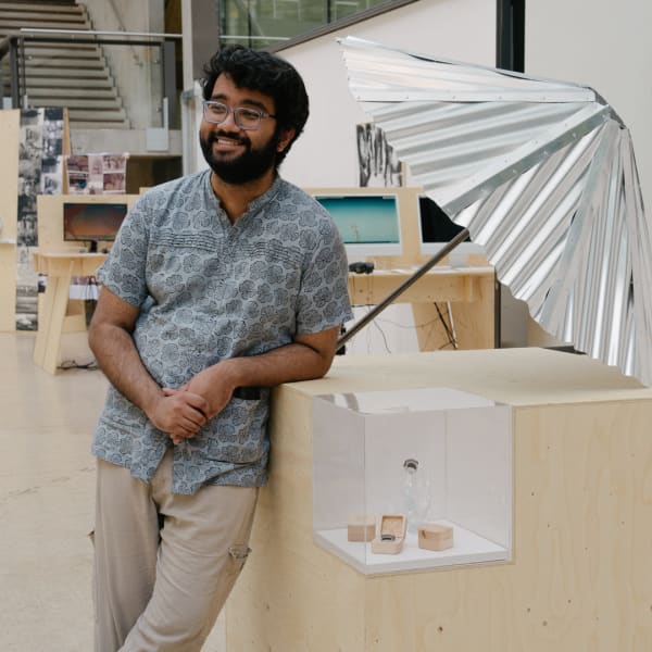 A student stands smiling in a studio, leaning against a wooden display plinth with a metal umbrella installation on top, surrounded by desks and creative materials.