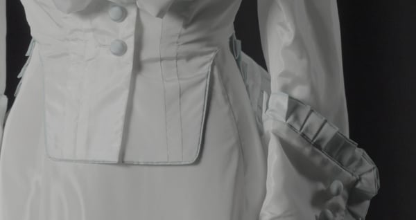 Close-up of a white, satin-like dress featuring structured seams, large buttons, and intricate ruffled details on the sleeves, conveying elegance and sophistication.