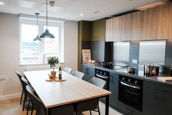 A shared kitchen and dining area with dark cabinetry, a black induction hob, oven, stainless steel splashback, and a large wooden dining table with six grey chairs, lit by two industrial-style pendant lights above.
