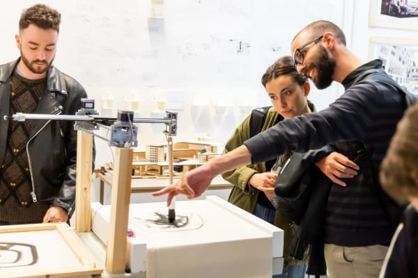 A drawing machine is creating a piece of work in a room. man in glasses with a shaved head and beard is talking to a women who leans in to listen to him as he gestures at it. To the left, a man in a leather jacket is looking down at it as it works.