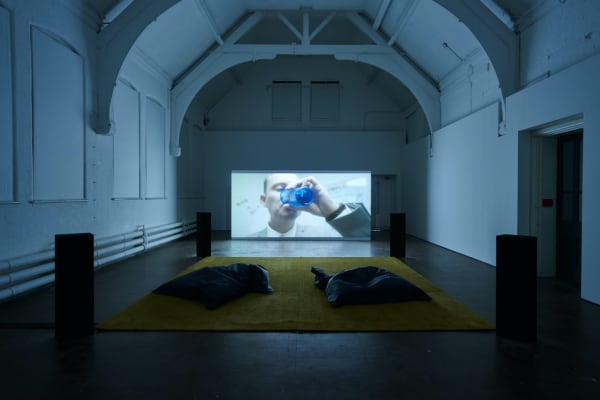 Dark screening room with cushions on the floor and an official man in a suit drinking water on screen