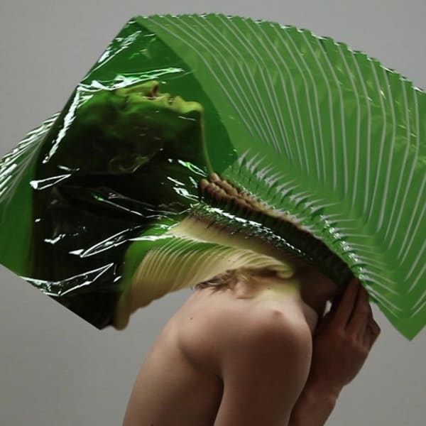 Distorted image of topless model, body leaning forward, head leaning back within a large plastic green leaf textile.