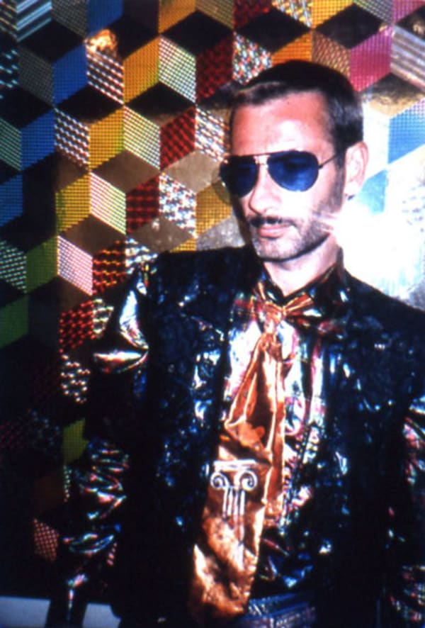 David McDiarmid posed in front of a multicoloured, holographic geometric patterned wall. David wears a metalic black shirt and bronze tie, with navy sunglasses.