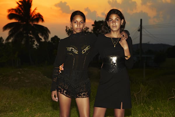 two models side-hugging in black dress garments, posed outdoors in front of a sunset landscape