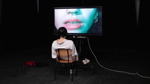 Girl watching a video installation with a close up of a girls mouth, pierced nose and ear on the screen. Work by Eva Mateos Rodriguez - BA Contemporary Theatre and Performance