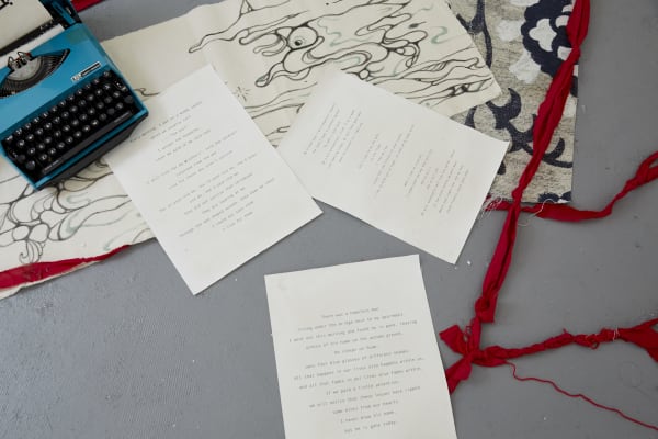 A blue typewriter is in the corner, with three pages of paper with poems. There are red ribbons and textiles across the floor