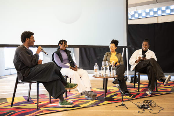 Four people sit in a line in front of an audience, with microphones in their hands. One is speaking, and the others smiling and laughing.