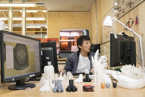 A student seated at a table with a monitor, lamp and lots of small models and prototypes of different shapes and sizes.