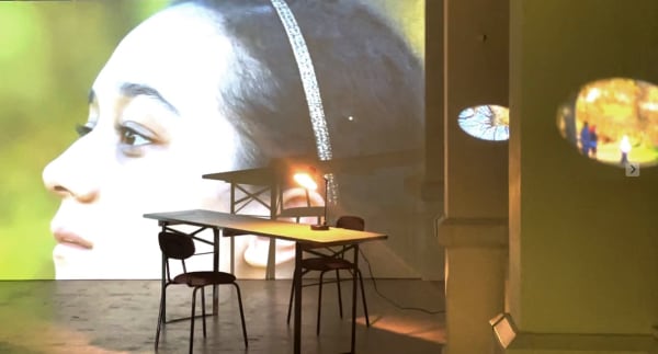 A table and chairs next to a wide screen with a person's face projected. Other light effects are visible on the image, suggesting an exhibition installation.