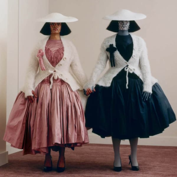 A square shot of 2 models in similar garments, holding hands, wearing hats and cardigans. One appears in colour and the other in a black and white version of the same clothing.